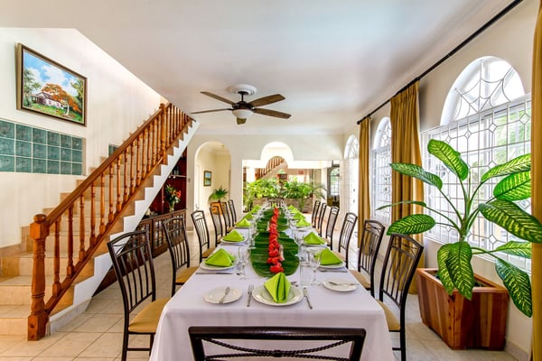 Formal dining room with long table set for dinner, black chairs, green napkins, wooden staircase, arched windows with gold curtains, and potted plants.