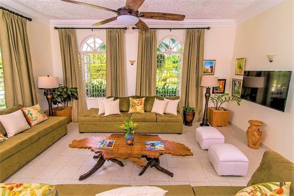 Spacious living room with olive-green sofas, wooden coffee table, arched windows overlooking garden, ceiling fan, and decorative plants
