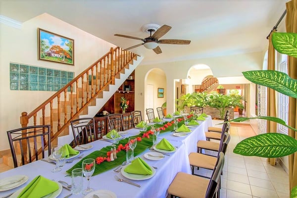 Formal dining room with long table set for event, wooden staircase, ceiling fan, potted plants, and arched doorways leading to adjacent spaces