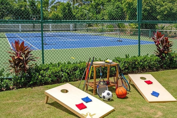 Fenced tennis court with cornhole boards, sports equipment, and red flowering plants on grass lawn