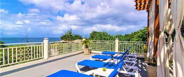 Oceanfront terrace with blue lounge chairs, white railing, pergola, and tropical ocean view under cloudy sky