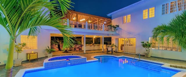 Two-story white house with illuminated pool and spa at dusk, palm trees, wooden deck, and upper balcony.