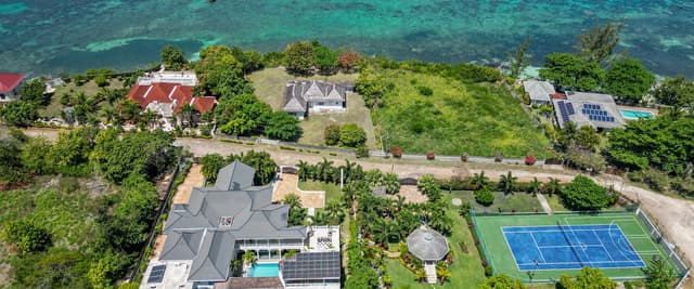 Aerial view of beachfront properties with turquoise water, including houses with solar panels, swimming pool, and tennis court