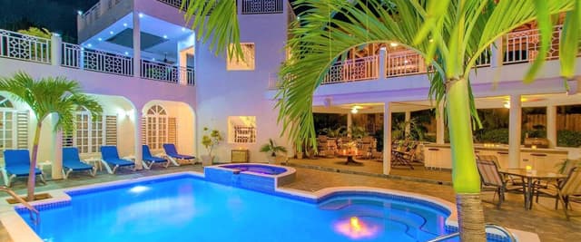 Illuminated pool and hot tub at night with pink villa, palm trees, lounge chairs, and covered terrace