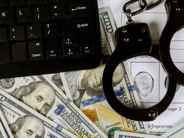 Black keyboard, handcuffs, US dollar bills, and fingerprint chart on white surface