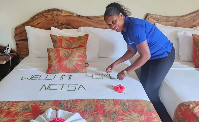 Staff member arranging welcome message and decorative towel on bed with wooden headboard and patterned bedding