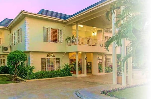 Two-story cream-colored house with dark roof, covered porch, driveway, and tropical palm trees at dusk
