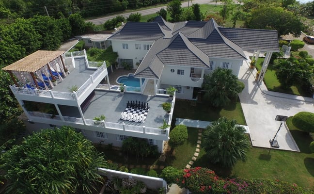 Aerial view of waterfront home with dark roof, pool, deck, manicured lawn, and water visible beyond tree-lined road.