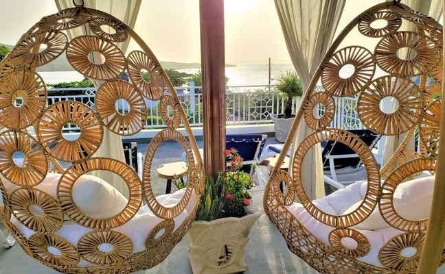 Two woven rattan hanging chairs with white cushions on a waterfront terrace, overlooking a bay with boats and distant shoreline.