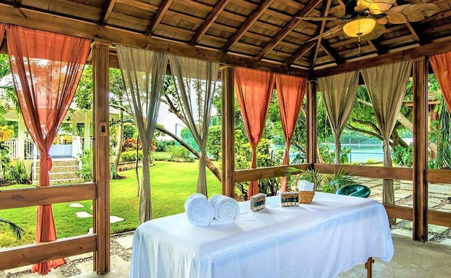 Covered outdoor pavilion with wooden beams, orange and tan curtains, massage table with rolled towels, overlooking manicured lawn and water view