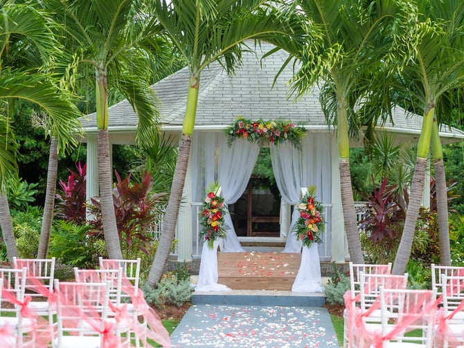 Outdoor wedding ceremony setup with pink chairs, palm trees, white draped archway, and floral arrangements