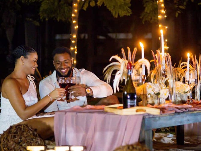 Couple toasting with wine glasses at outdoor nighttime dinner table with string lights and neon sign