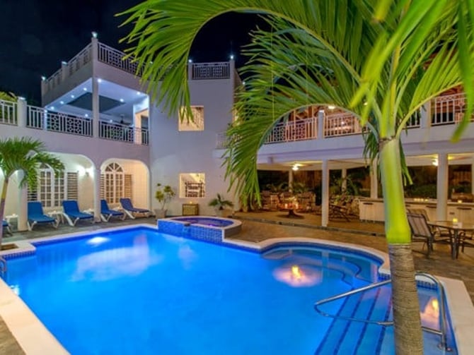 Illuminated swimming pool with spa surrounded by white villa with arched windows and palm trees at dusk