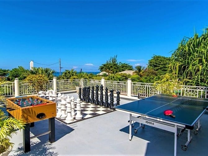 Elevated outdoor recreation deck with foosball table, giant chess set, and ping-pong table overlooking tropical landscape and ocean