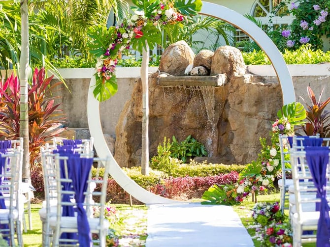 Tropical garden wedding ceremony setup with white arch, blue chairs, colorful flowers, and stone water feature