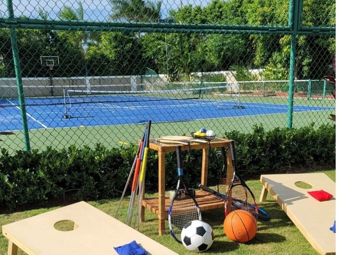 Fenced sports facility with blue tennis court, cornhole boards, and equipment including soccer ball, basketball, and tennis rackets