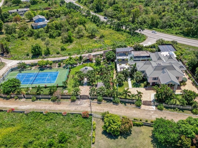 Aerial view of modern house with metal roof, blue tennis court, and manicured gardens surrounded by dense trees and rural road.