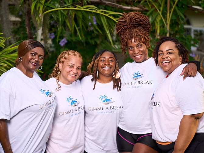 Five women wearing white Gimme a Break Retreats t-shirts posing together in a tropical garden setting with palm fronds and purple flowers in background.