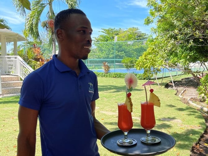 Staff member holding tray with two tropical cocktails in lush backyard garden with tennis court and palm trees