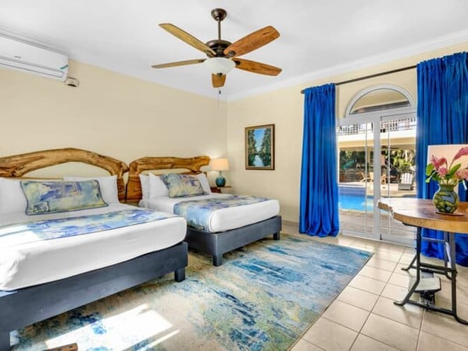 Bedroom with two beds, wooden headboards, ceiling fan, blue curtains, and glass doors opening to pool area