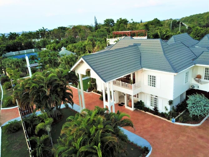 White two-story house with blue-gray roof, red tile driveway, tropical landscaping, and pool visible in lush hillside setting.