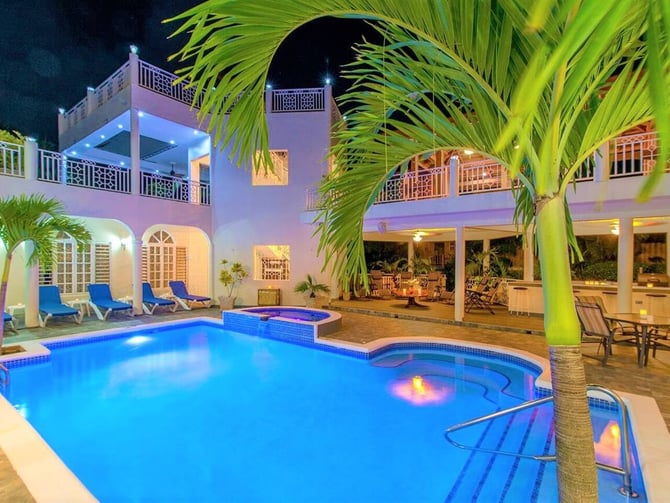 Nighttime view of pink villa with illuminated pool, hot tub, lounge chairs, and palm trees