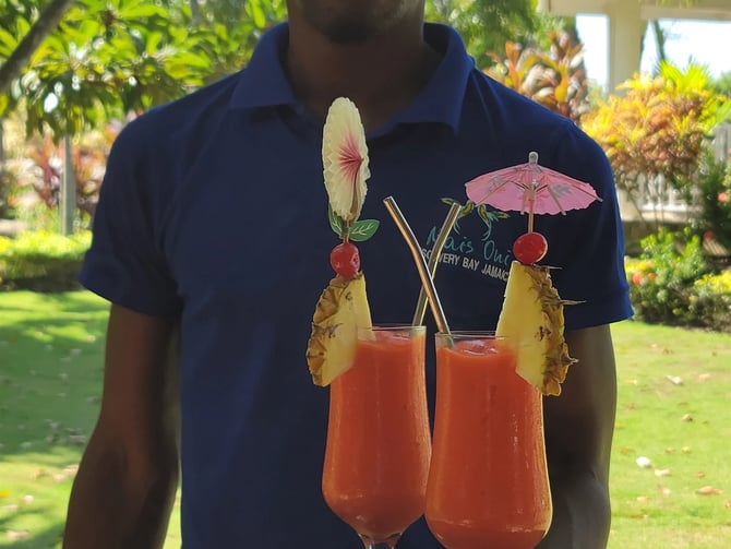 Man in blue polo shirt holding tray with two tropical cocktails garnished with pineapple, flower, and umbrella in lush garden setting