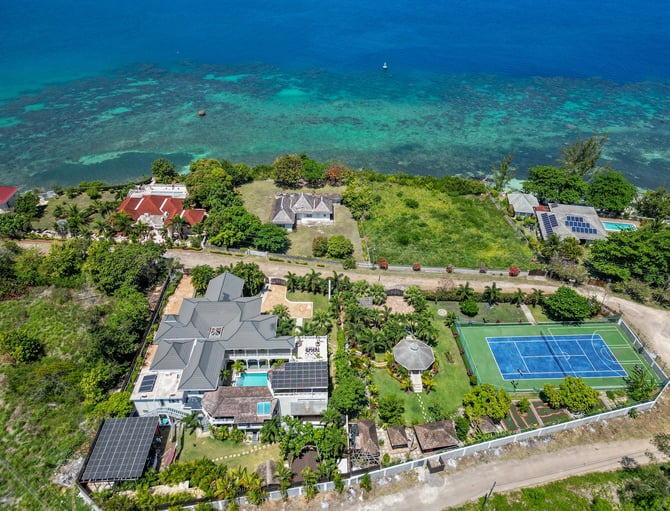Aerial view of beachfront properties with turquoise water, including homes with pools, tennis court, and solar panels